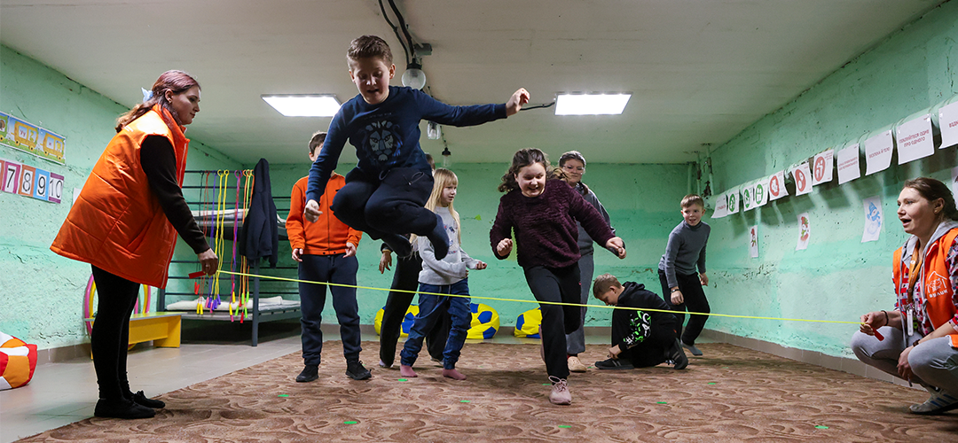 Blick in einen Schutz- und Spielraum in Sumy: Hier lernen Kinder mit Stress umzugehen, Resilienz aufzubauen und wieder zu sich selbst und anderen zu finden. © Iryna Prytchyna / Save the Children Mehrere Kinder, die über ein Seil springen, das von zwei erwachsenen Personen gehalten wird.