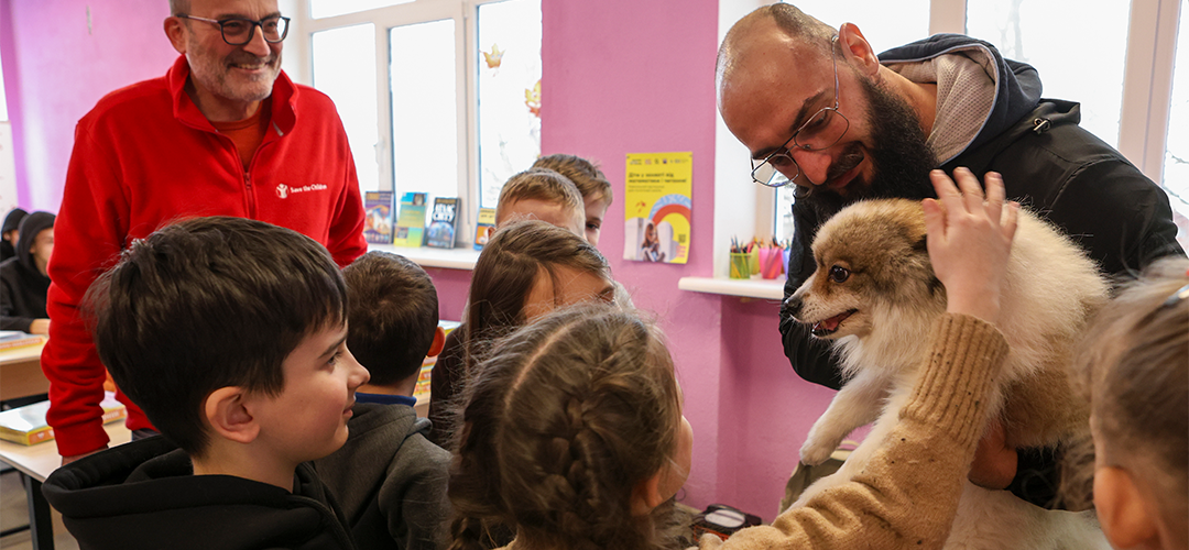 Für viele Kinder ist der Minenspürhund das Highlight der Gefahrenschulung. © Iryna Prytchyna / Save the Children Eine erwachsene Person hat einen Hund auf dem Arm. Mehrere Kinder stehen drumherum und streicheln den Hund.