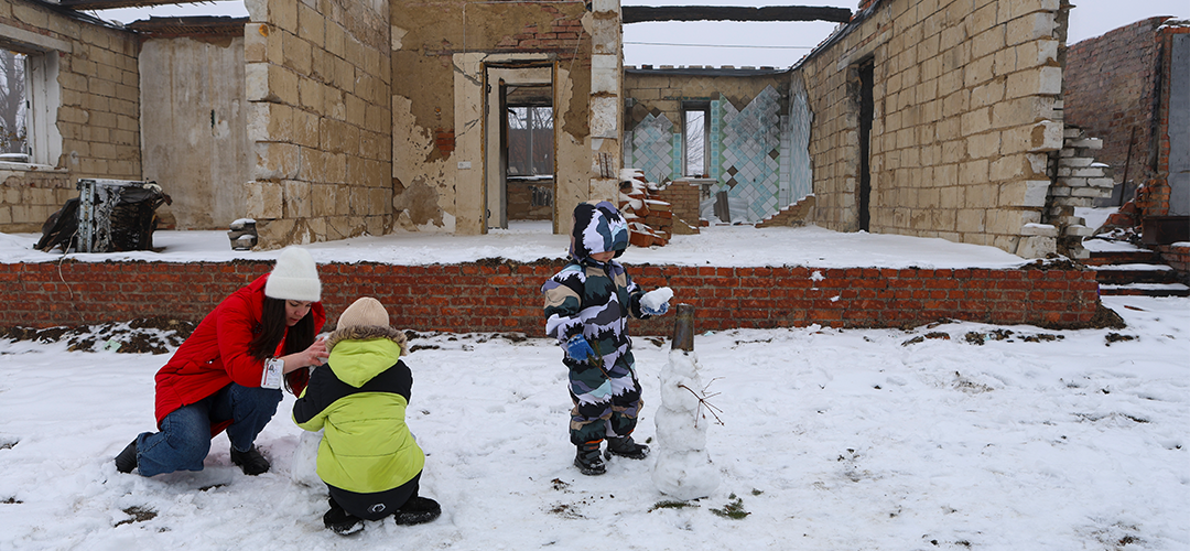 Vor den Ruinen ihres zerstörten Hauses spielen die Kinder im Schnee. © Iryna Prytchyna / Save the Children Zwei Kinder und eine erwachsene Person bauen einen Schneemann. Hinter ihnen die Ruinen eines Hauses.
