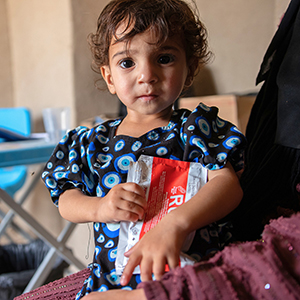 Nahrung statt Not: Ein Kleinkind hält Spezialnahrung in einem Raum mit Tisch und Stühlen. © Sacha Myers / Save the Children Ein Kleinkind hält Spezialnahrung in einem Raum mit Tisch und Stühlen.
