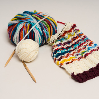 Die Aktion „Ein Schal fürs Leben“ von Brigitte und Save the Children lädt auch 2026 zum Stricken für den guten Zweck ein. © Save the Children Zwei Wollknäuel, der Anfang eines gestrickten Schals und Stricknadeln liegen auf einem Tisch.